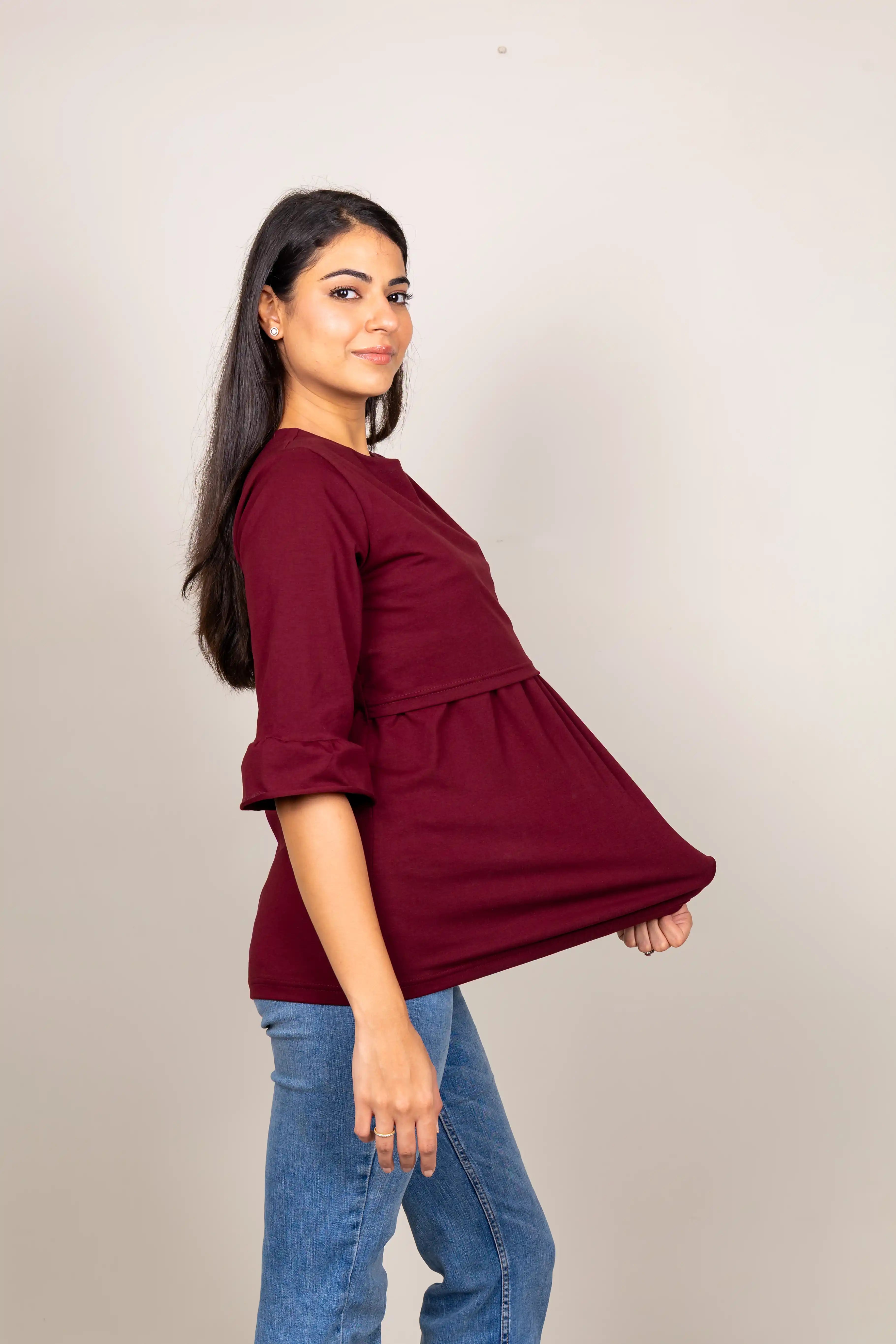 A woman modeling a three-quarter sleeve peplum top in deep maroon, paired with blue jeans, against a plain light gray studio background.