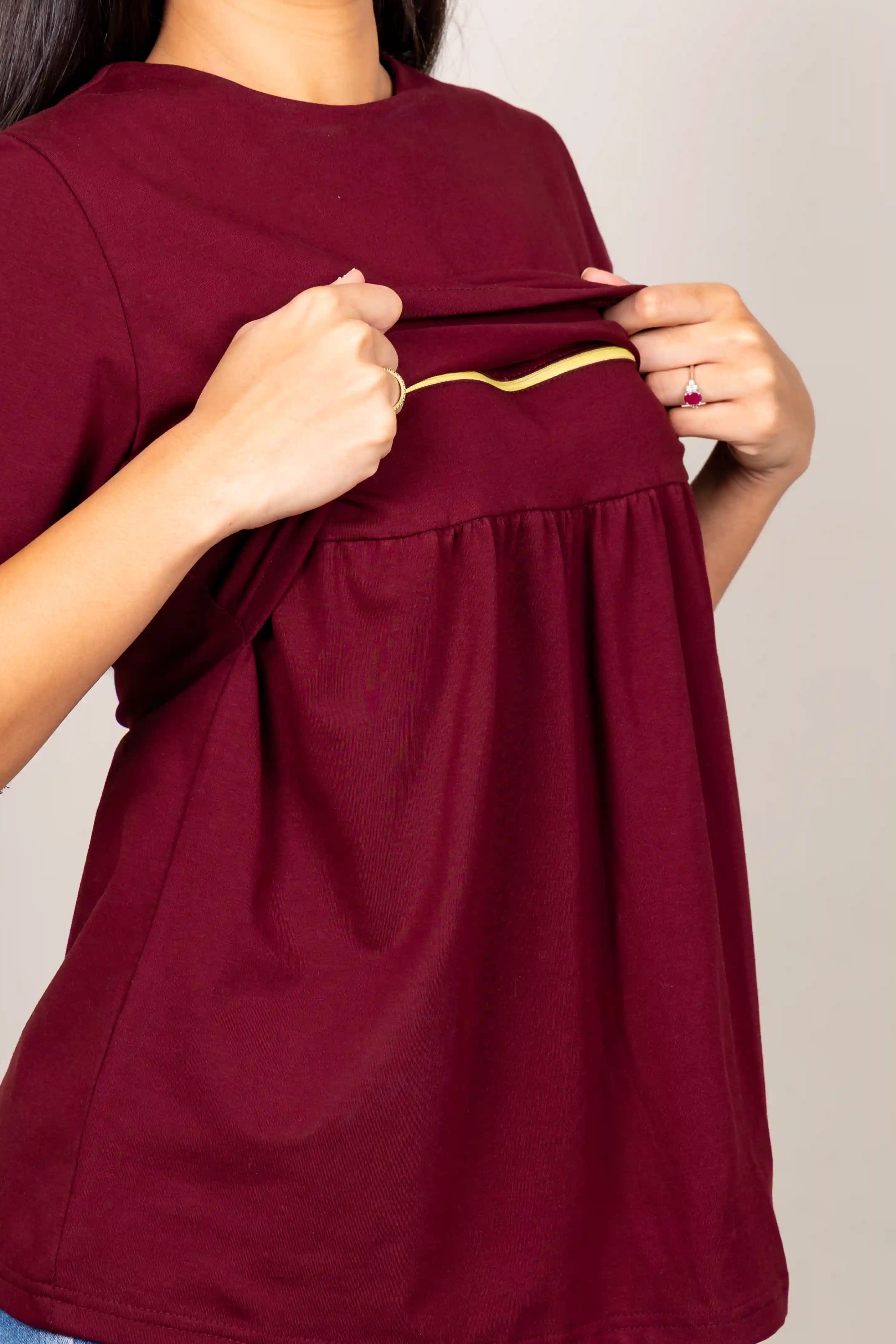 A woman wearing a short-sleeve burgundy maternity top, holding up the front elastic band to show the stretch. The top has a gathered waist and is paired with blue jeans against a plain light gray background.