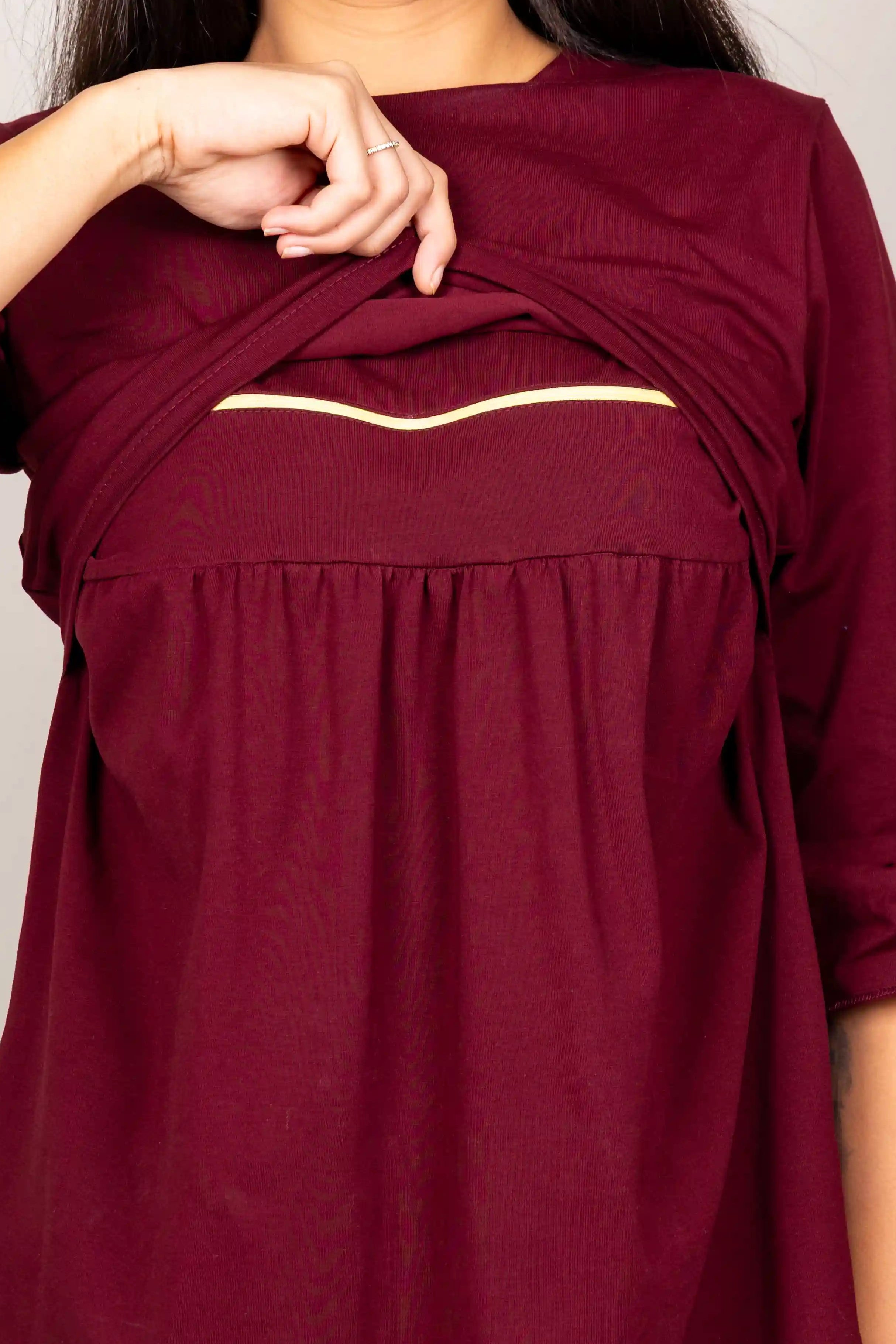 A woman wearing a burgundy maternity top with a built-in front pouch pocket, lifting the fabric to show the pocket. The top has three-quarter sleeves and a relaxed fit, paired with blue jeans against a plain light gray background.