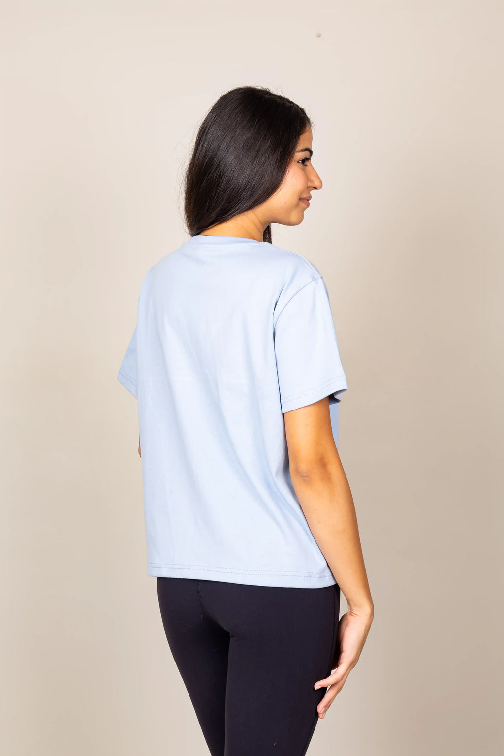 A woman shown from the back wearing a light blue short-sleeve t-shirt and dark leggings, standing against a plain beige studio background.