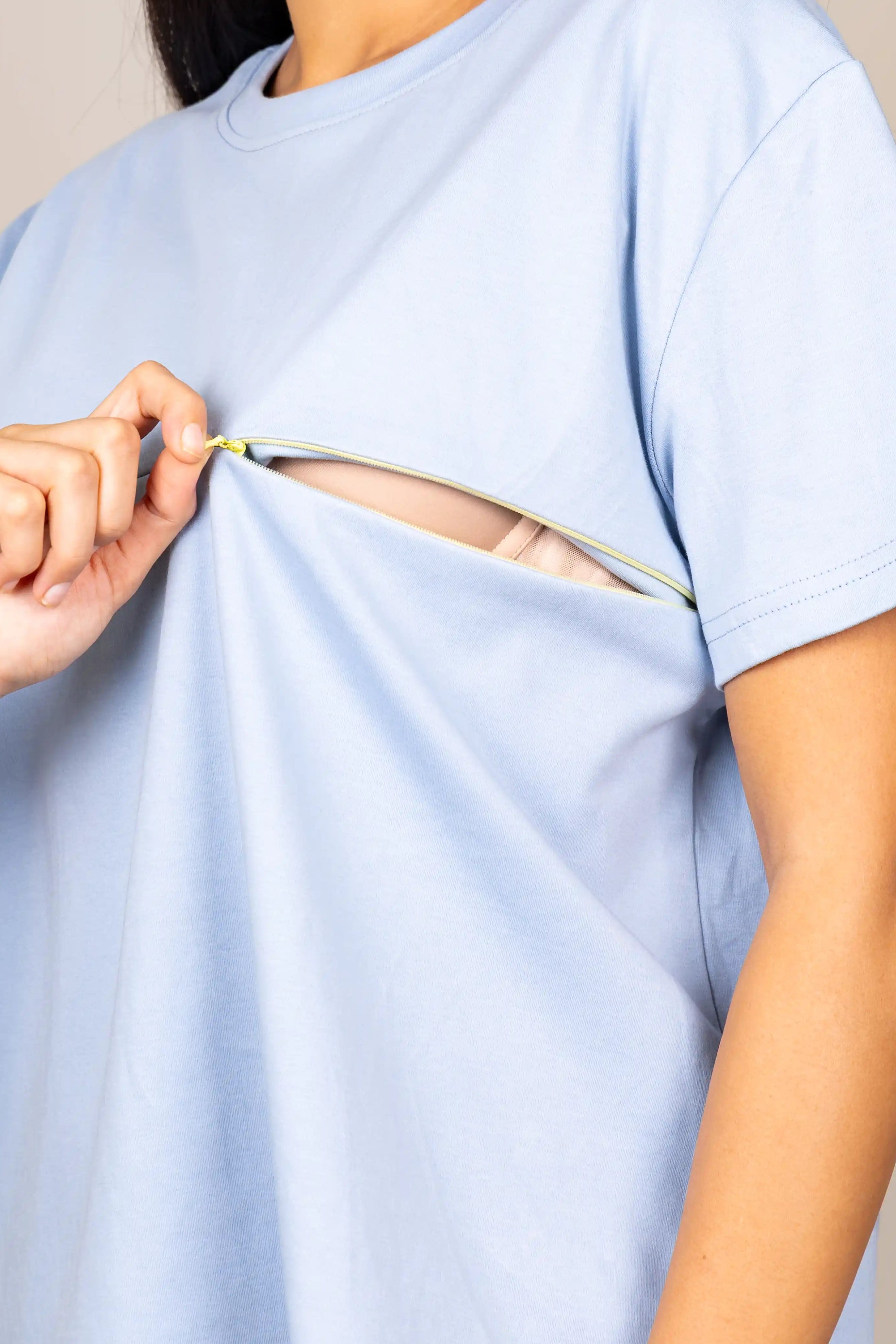 A woman wearing a light blue short-sleeve t-shirt with a horizontal zipper cutout across the chest, showing a zipper pull and a hint of skin underneath. She is holding the zipper tab with her hand against a plain beige background.