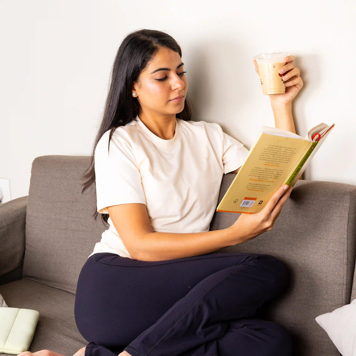 A woman sits cross-legged on a gray sofa, reading a book and holding a takeaway coffee cup. She is wearing a white t-shirt and dark navy lounge pants.